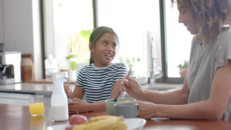 Happy-biracial-mother-and-daughter-eating-breakfast-cereal-talking-in-sunny-kitchen,-slow-motion