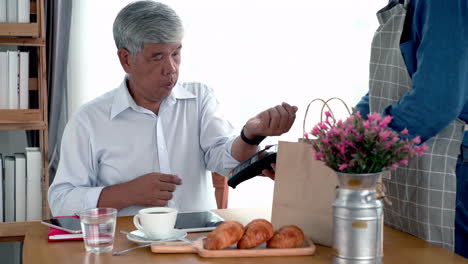 active senior asian man working in a coffeeshop using a mobile device, smartwatch for nfc, qr code scanning, contactless payment to pay for coffee and snack