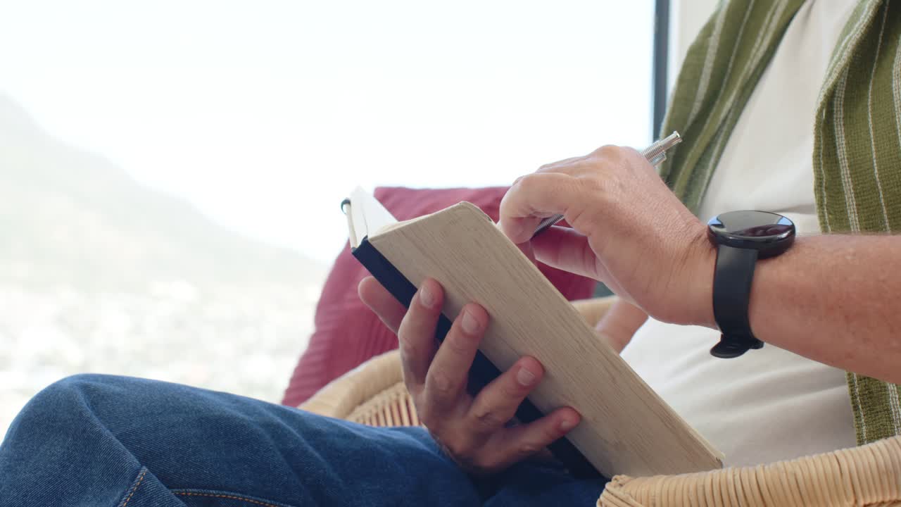 Seated man is tapping pen and is taking notes in notebook beside bright window, showing wristwatch