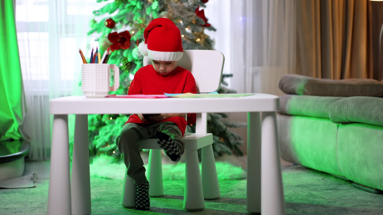 Child in Christmas hat coloring at home. A young child wearing a festive hat sits at a table coloring, with a decorated Christmas tree in the background