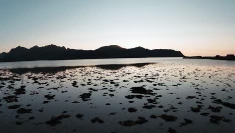 Fliegen-Am-Ufer-In-Bodennähe-Mit-Blick-Auf-Das-Seichte-Wasser,-Die-Berge-Im-Hintergrund-Und-Die-Untergehende-Sonne-Dahinter