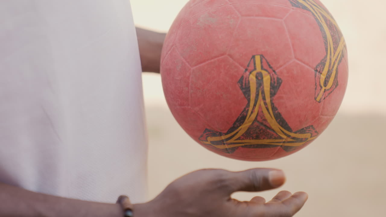 Ação de futebol de praia ao pôr do sol. Jogador sereno manobra habilmente a bola carmesim no cenário do pôr do sol. Atleta focado controla e gira com perícia a bola de futebol vermelha texturizada em meio à cena tranquila da praia.