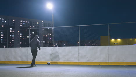 vista trasera de un atleta con capucha jugando al fútbol por la noche bajo la iluminación brillante del estadio, con una pelota de fútbol cerca de la pared que refleja la silueta, el paisaje urbano en el fondo