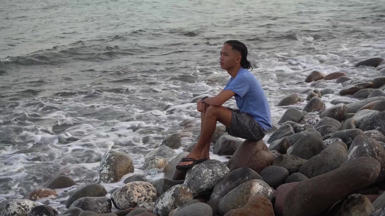 Asiatisk filippinsk ung dreng sidder alene ved en klippefyldt kyst ved Town Rock Beach i San Juan-provinsen, Leyte, Filippinerne. Statisk optagelse.