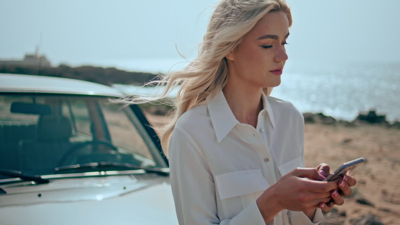 Splendida modella usa lo smartphone sulla spiaggia oceanica, appoggiata a un'auto d'epoca, primo piano.