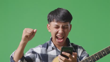 스마트 폰을 사용하여 기타를 들고 녹색 화면 배경에 서서 성공을 축하하는 아시아 십대