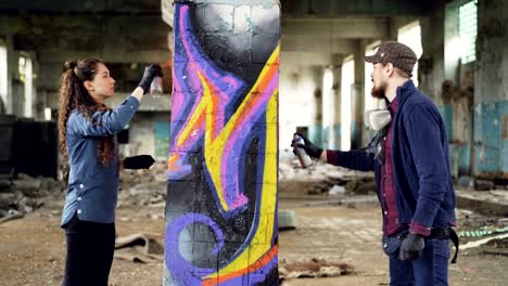 concentrated man and woman team of urban painters are painting graffiti on pillar inside ruined empty house using aerosol paint. dirty interior in background.