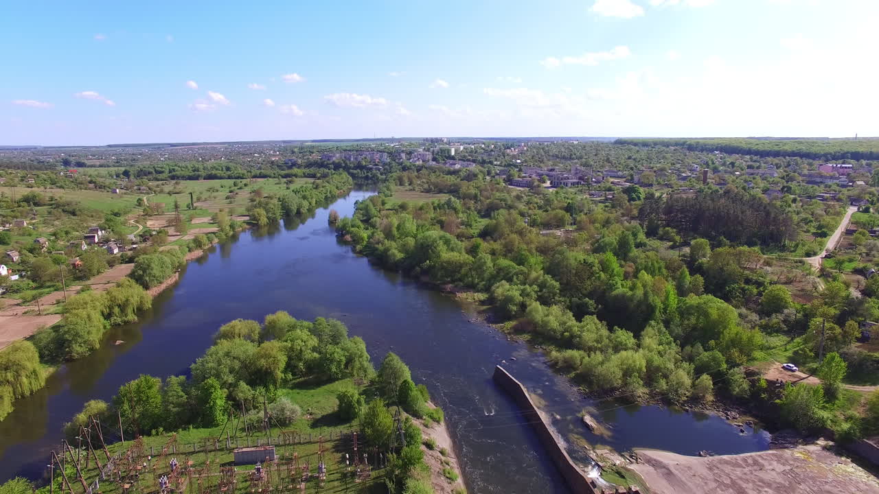 Bridge and dam over the narrow river. Drone footage over the river flowing between picturesque banks with lots of greenery. City scenery at backdrop.