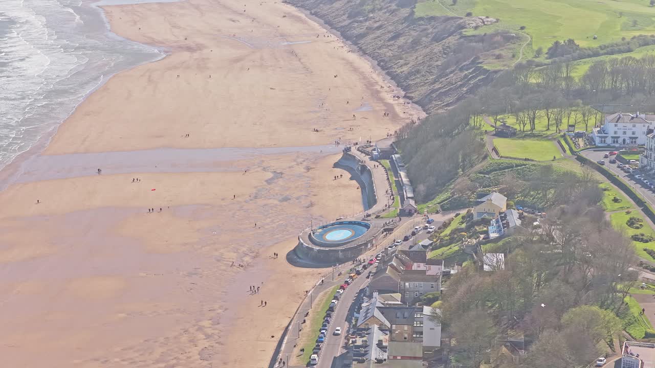 Piscina naturale di Filey Promenade su Ravine Road, Filey, North Yorkshire, Inghilterra, con piscina circolare di acqua di mare, muro frangiflutti curvo e ampia spiaggia sabbiosa lungo la costa del Mare del Nord, ripresa orbitale con drone