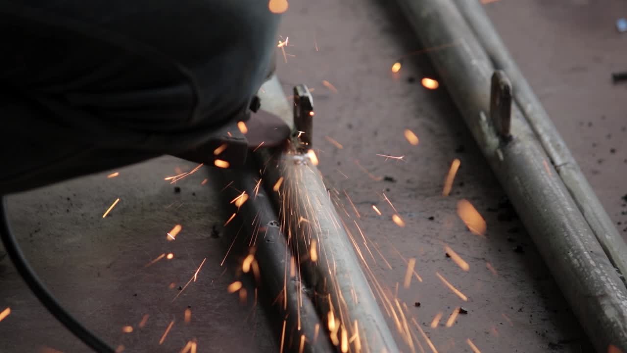 Rimozione dell'eccesso di saldatura da tubi di ferro per una finitura liscia, con smerigliatrice elettrica manuale da parte di un tuttofare, clip in slow-motion all'aperto, con scintille di particelle che vanno verso la telecamera.