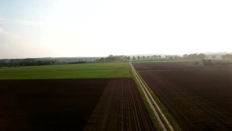 Aerial-view-of-large-farmland-being-cultivated-and-harvested-on-a-clear-summer-day,-panning-to-showcase-the-wide-farmland-on-rural-countryside-with-misty-background