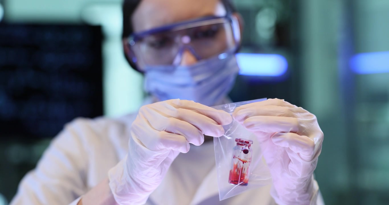 Technician puts vial with red liquid into plastic bag in lab