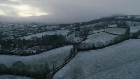 Monotone-looking-drone-shot-of-a-wintry-snowy-scene-in-east-Devon-farmland-following-a-country-road-and-a-car-driving-in-icy-conditions