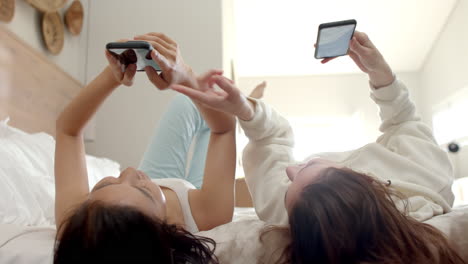 lying on bed, two diverse women friends using smartphones and relaxing at home