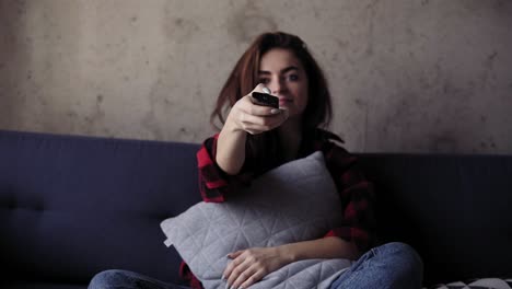 young beautiful brunete girl is holding a pillow while sitting on sofa. she tries to turn the tv on but the remone control is not working.