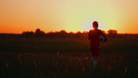 el niño está en el campo al atardecer en la hierba con una pelota de fútbol. soñando con una carrera futbolística. el concepto de éxito en el deporte.