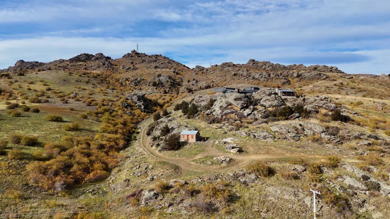 Drone footage captures a solitary cabin amidst rocky hills and sparse vegetation under clear skies in Cromwell, New Zealand