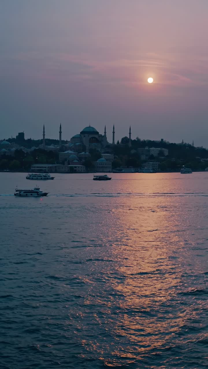 Ferries are sailing on the calm sea at sunset, with a mosque and minarets silhouetted against the colorful sky, creating a serene and picturesque scene