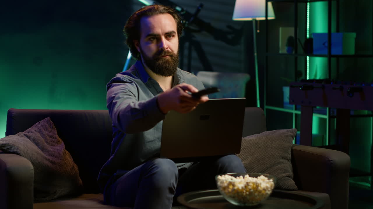 Uomo che digita al laptop, seduto sul divano e guarda la TV.