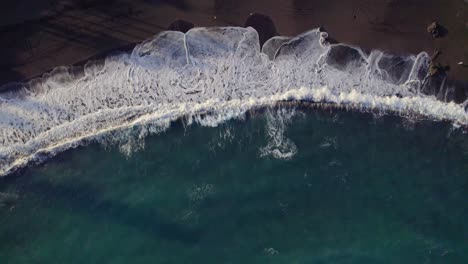Top-down-drone-view,-turquoise-waves-breaking-on-black-sand-beach