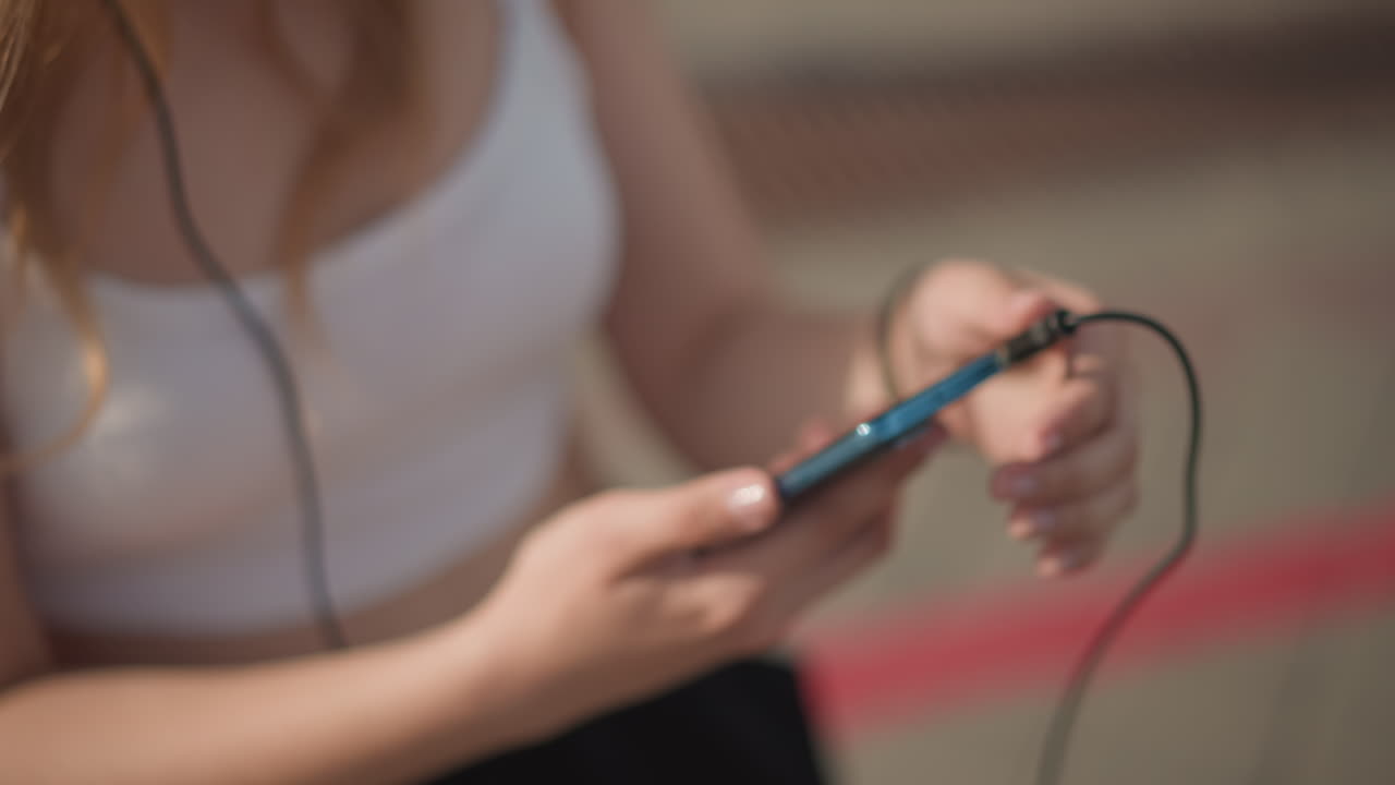 white female on street putting on headphones, plugging cable into smartphone and testing audio, fingers adjusting cord while midriff shows in crop top and black pants, hair catching sunlight, calm