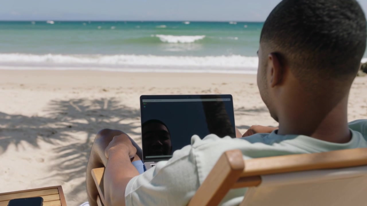 Starting relaxed, man wearing tee and shorts working on laptop at beach, smartphone on side table