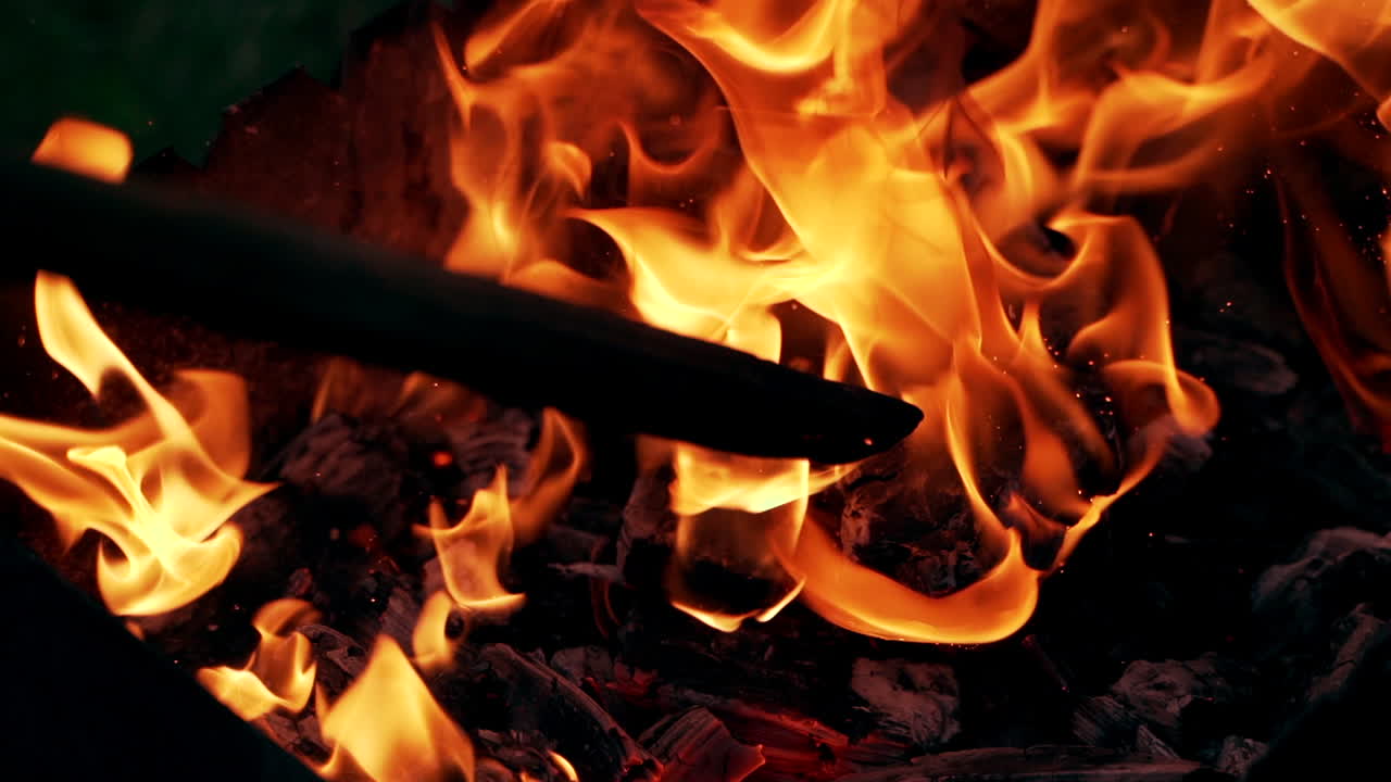 Bright fire motion. Sparkles of red and yellow flame from smoldered logs. Wooden stick beats a black log in the burning fire. Slow motion.