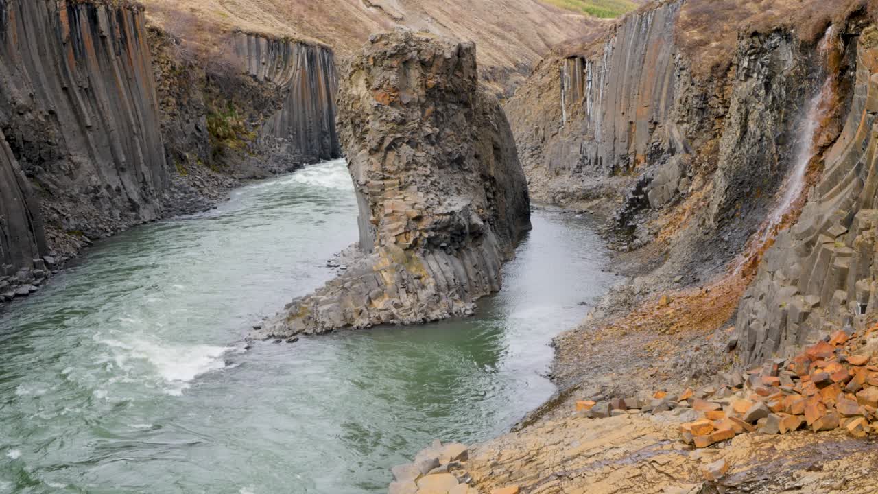 Rushing river flows through a dramatic Icelandic canyon with distinctive basalt column formations and rugged terrain, showcasing nature's power