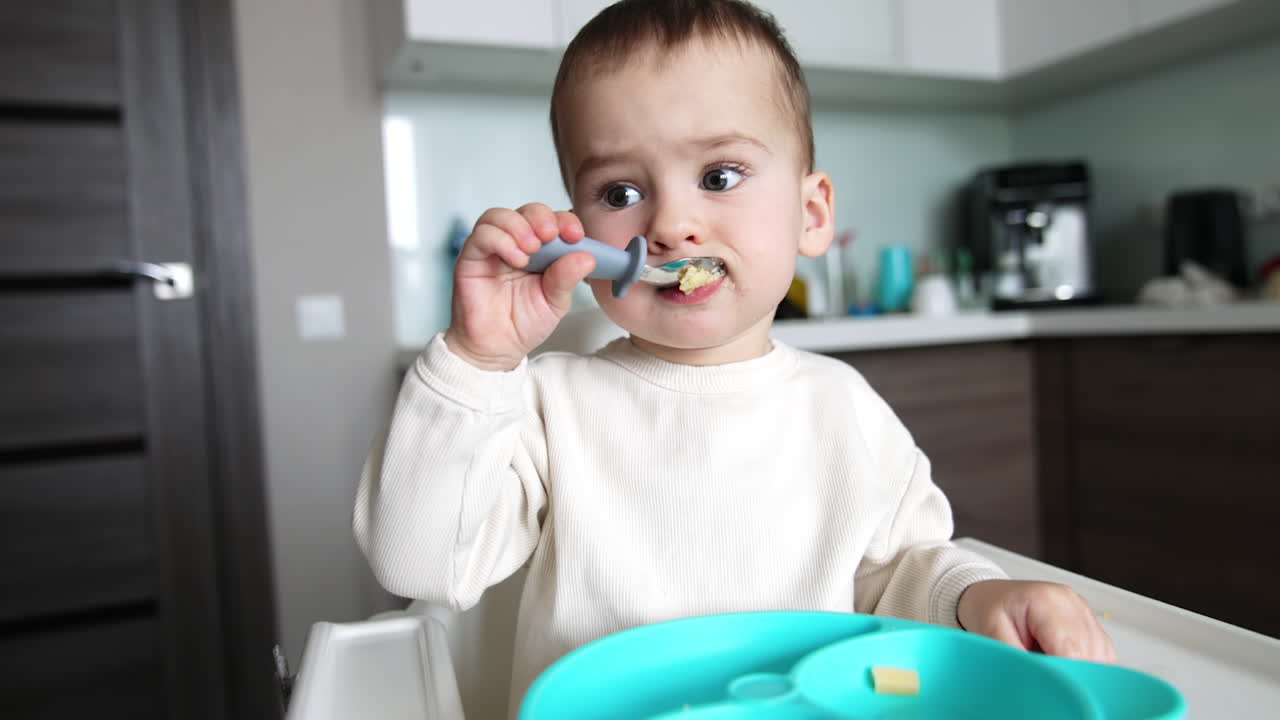 Small baby boy eating food from spoon himself. Charming kid has some porridge for breakfast. Close up.
