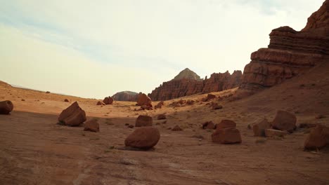 A-gorgeous-tilt-up-shot-of-a-stunning-desert-plane-with-large-rocks-scattered-across-the-ground-with-large-red-rock-mountains-on-the-right-on-a-small-hike-in-Utah-State-Park,-Goblin-Valley