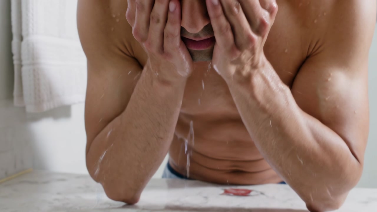 Faucet running, shirtless man cupping, washing and drying face with white towel at bathroom sink