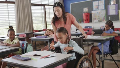 maestra diversa que enseña a una escolar en silla de ruedas que lee en el aula de la escuela primaria