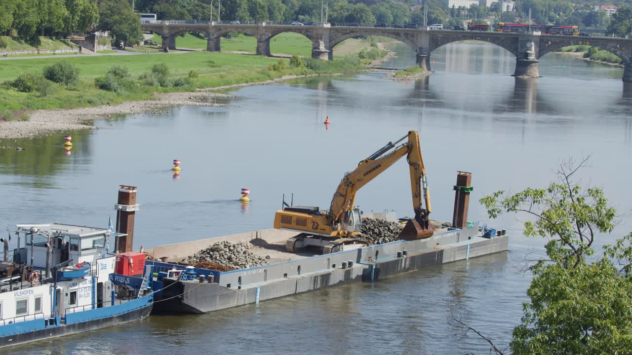A large excavator transfers rocks onto a barge in a river, with bridges and greenery in the background. Daylight, wide aerial view, steady camera