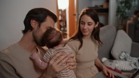 Happy-brunette-man-with-stubble-holds-his-little-baby-daughter-in-his-arms-along-with-his-young-wife-in-a-modern-apartment-on-a-gray-sofa