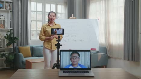 asian female teacher shoots video by smartphone camera and having video call on laptop while teaching math at home