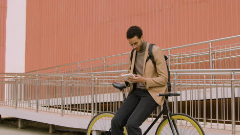 young american man in formal clothes using a tablet while leaning on bike in front of a prefab metal building