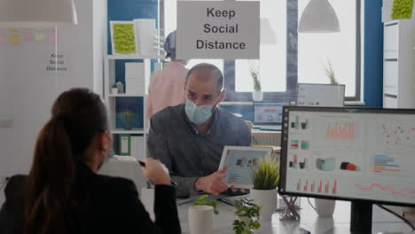 Group-of-business-coworkers-with-face-masks-analyzing-graphs-using-digital-tablet