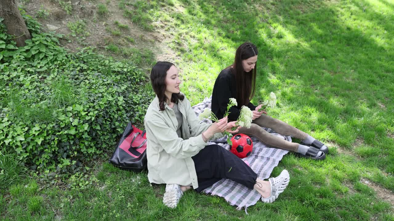 Para wanita duduk di selimut menikmati aroma lembut bunga kamomil di taman.