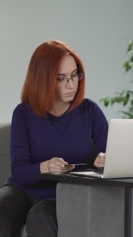 red-haired woman in glasses enters credit card details on website via laptop. young housewife makes online payment sitting on sofa in living room at home