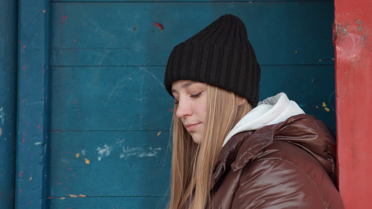 도시의 고독을 느끼며 생각에 잠긴 코트를 입은 소녀가 파란색 배경 앞에서 내성적인 모습을 보입니다. 고풍스러운 옷차림의 한 여성이 도시 풍경의 벽에 기대어 앉아 경계심 있는 내면의 성찰을 표현합니다.