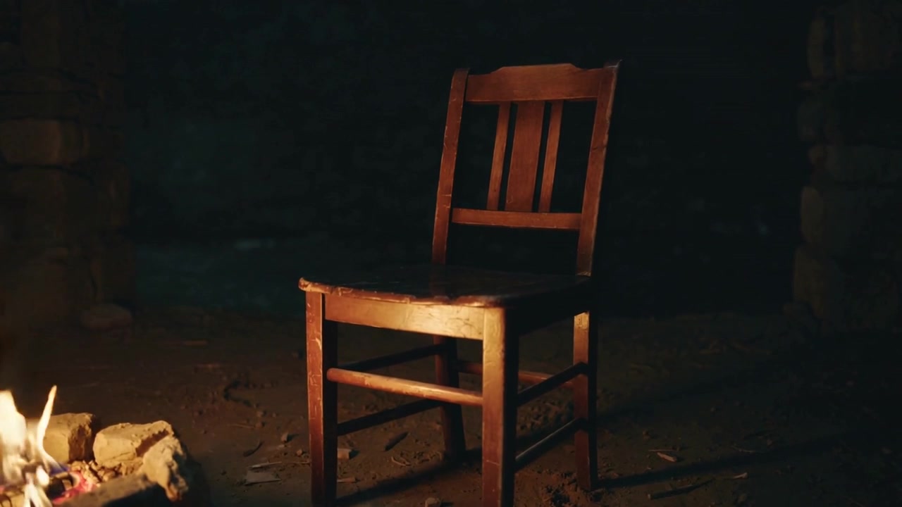 Pulzující plameny z kamenného ohniště osvětlují dřevěnou židli na podlaze chaty a zvýrazňují opotřebované sedadlo.