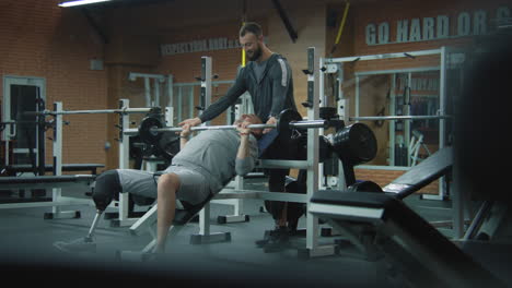 Athletic-man-with-prosthetic-leg-performs-weight-training-with-barbell-in-gym-with-fitness-therapy-trainer,-then-takes-a-break.-Sportsman-with-artificial-limb-does-exercises-using-sports-equipment.