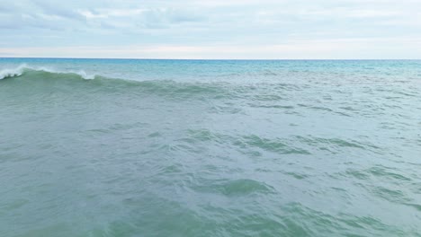 Aerial-dolly-above-green-Caribbean-water-as-gentle-ocean-wave-crashes,-blue-horizon