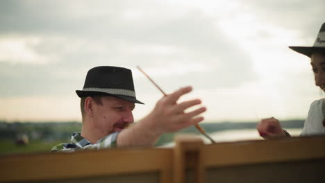 a painter in a hat and checkered shirt is focused on his work as a woman in a white dress and hat playfully touches his nose with a blade of grass, sharing a light-hearted moment outdoors