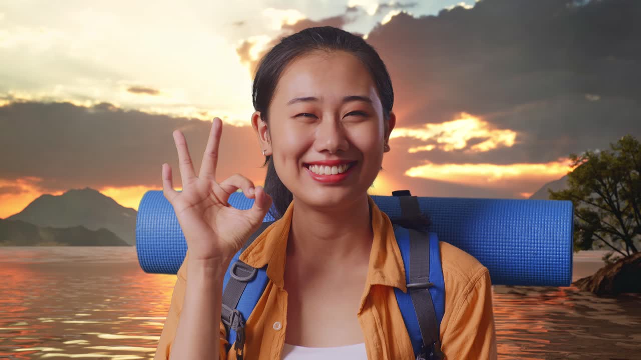 Close Up Of Asian Female Hiker With Mountaineering Backpack Smiling And Showing Okay Gesture To Camera While Standing At A Lake During Sunset Time