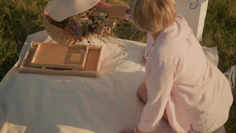 lady in pink shirt sitting on picnic mat, reaching for basket containing flowers and sun hat to keep close to her leg, white paper bag nearby, warm sunlight