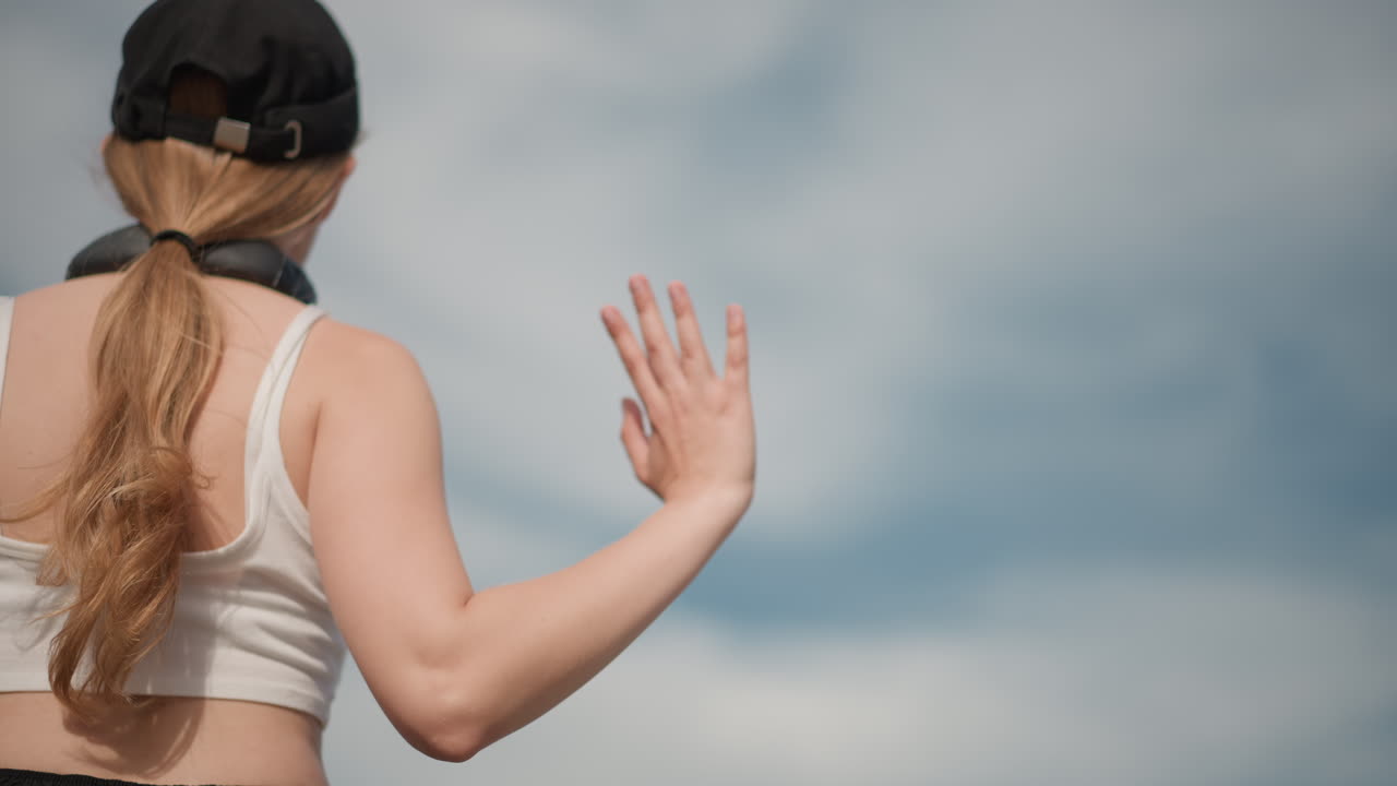 Ballerina vista da dietro all'aperto che allunga le braccia verso il cielo, top corto bianco, cappellino nero, cuffie, lunga coda di cavallo, ampi gesti con le braccia, movimento freestyle fluido, luce del giorno intensa, cielo azzurro nuvoloso, prova casual in strada