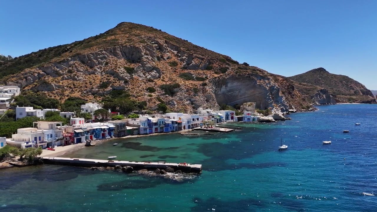 Il villaggio di pescatori di Klima costeggia la costa turchese con colorate rimesse per barche syrmata sotto aspre scogliere vulcaniche, affacciato sul Mar Egeo a Milos, Grecia, scivolata laterale del drone verso la costa.