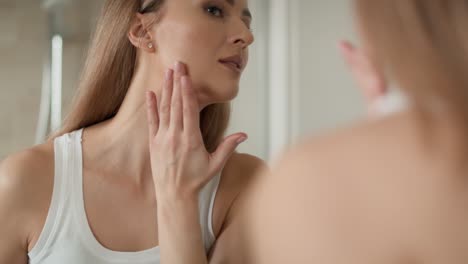 joven mujer caucásica sonriendo y aplicando crema facial en el reflejo del espejo.