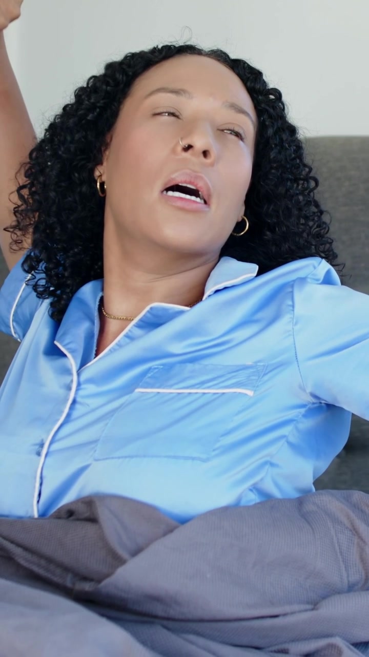 Vertical video: Waking woman in blue satin pajamas sitting up on bed, pulling blanket to start day